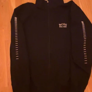 Svart tröja från Hugo Boss - Snygg svart tröja från Hugo Boss med dragkedja och vita detaljer på ärmarna. Perfekt för en stilren look. Tröjan har långa ärmar och en hög krage.