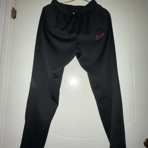 Svarta träningsbyxor från Nike - Snygga svarta träningsbyxor från Nike med röd broderad logga. Byxorna har en elastisk midja och är tillverkade i polyester för optimal komfort och rörelsefrihet. Perfekta för träning eller en avslappnad dag.