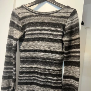 Grå och svart randig stickad tröja - Säljer en mysig stickad klänning från Nelly i grått och svart med randigt mönster. Klänningen har långa ärmar och en rund halsringning. Perfekt för kyligare dagar! Kort klänning 