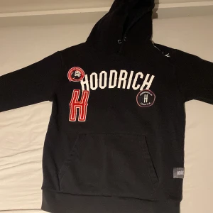 Svart hoodie från Hoodrich - Säljer en svart hoodie från Hoodrich med tryck och broderade detaljer i vitt och rött. Den har en stor ficka framtill och en justerbar huva. Perfekt för en avslappnad stil.