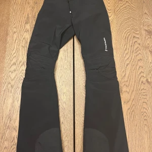 Svart skidbyxa från Peak Performance - Snygga svarta byxor från Peak Performance med dragkedja och knappar i midjan. Byxorna har en stilren design och är perfekta för utomhusaktiviteter. De är tillverkade i ett slitstarkt material som passar bra för kyligare väder. En knapp har lossnat uppe vid midjan, men det påvekrar inte hur dem sitter. 