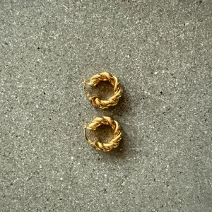 Guldiga  örhängen - Snygga guldiga örhängen med en unik ”flätad” design. Perfekta för att ge en elegant touch till din outfit. De är runda och har en fin glans som fångar ljuset vackert. Aldrig använda!