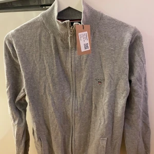Grå kofta från GANT - Säljer en stilren grå kofta från GANT i stretchig bomull. Koftan har en dragkedja framtill och en liten logotyp på bröstet. Perfekt för en avslappnad men snygg look. (Obs vet ej storleks nr, står S men passar även M)