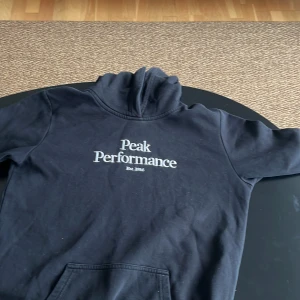 Svart hoodie från Peak Performance - Säljer en stilren svart hoodie från Peak Performance med tryck på bröstet. Perfekt för en avslappnad stil. Hoodien har en klassisk design med känguruficka och ribbade muddar.