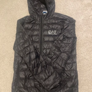 Svart pufferjacka från Emporio Armani - Säljer en svart pufferjacka från Emporio Armani med EA7-logga på bröstet. Jackan har en dragkedja framtill och en huva för extra skydd mot väder. Perfekt för kyligare dagar. Kom med prisförslag och säg vilken ni vill ha