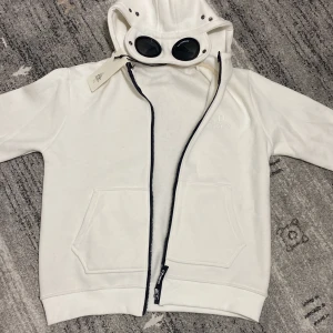 Vit huvtröja med glasögon från C.P. Company - Säljer en cool vit huvtröja från C.P. Company med inbyggda glasögon i huvan. Tröjan har dragkedja framtill och två fickor. Perfekt för en unik stil!