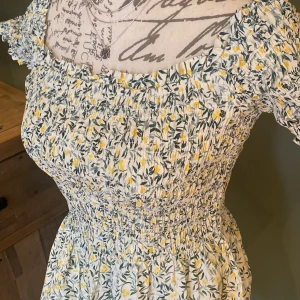 Blommig sommarklänning - Charmig blommig sommarklänning med korta ärmar och elastisk smockdetalj. Klänningen har ett mönster av små gula och vita blommor på en ljus bakgrund. Perfekt för varma dagar med sin luftiga design och bekväma passform.