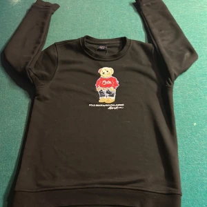Svart tröja med Polo Bear från Ralph Lauren - Säljer en svart tröja från Ralph Lauren med det ikoniska Polo Bear-motivet på framsidan. Tröjan har långa ärmar och en rund halsringning. Perfekt för en stilren och avslappnad look. Behövs bara tvättas men det löser jag