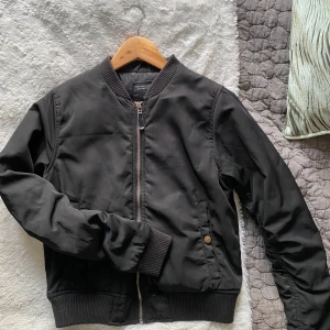 Svart bomberjacka från Zara - Snygg svart bomberjacka från Zara med dragkedja och ribbade muddar. Jackan har långa ärmar och två fickor med knappstängning. Perfekt för en cool och avslappnad stil.