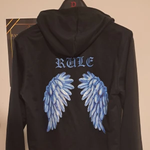 Svart hoodie med vingar - Säljer en svart hoodie med blå text 'RULE' på bröstet och stora blå vingar på ryggen. Tröjan har en klassisk huva med dragsko och långa ärmar. Perfekt för en cool och avslappnad stil.
