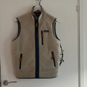 Beige fleeceväst från Patagonia - Säljer en snygg beige fleeceväst från Patagonia med blå dragkedjor och orange detaljer. Perfekt för lager-på-lager under kyliga dagar. Mörkt ljust på första bilden. 