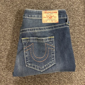 Blå jeans från True Religion - Snygga blå jeans från True Religion med klassisk femficksdesign och kontrastsömmar. De har en bootcut-stil och är perfekta för en avslappnad look. Märkets ikoniska logga syns på bakfickan. Använda ca 2 gånger 