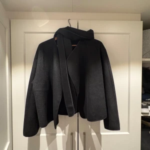 Svart tunn vårjacka med halsduk - Elegant svart kappa med matchande halsduk. Den har en stilren design med långa ärmar och en lös passform. Perfekt för kyligare dagar.