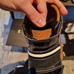 Svarta sneakers från Bally - Köpte för 5000 kronor. Snygga svarta sneakers från Bally med kardborreband och detaljer i brunt och vitt. Skorna har en hög modell med en robust sula och är tillverkade i skinn. Perfekta för en stilren look. 