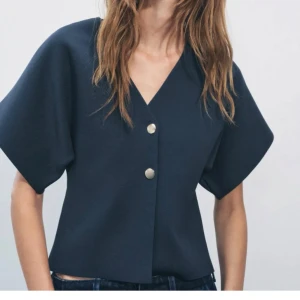 Mörkblå kortärmad blus från Zara - Snygg mörkblå blus från Zara med korta ärmar och v-ringning. Blusen har två dekorativa knappar framtill och en stilren design som passar perfekt till både jeans och kjol.