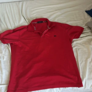 Röd pikétröja från Burberry - Säljer en snygg röd pikétröja från Burberry med klassisk krage och korta ärmar. Tröjan har ett diskret broderat märke på bröstet och ett stilrent rutigt mönster på insidan av kragen. Perfekt för en stilren look! Storlek är XL men den sitter som en medium-large. Har fått den av min pappa som säger att den är köpt i stockholm i butik.