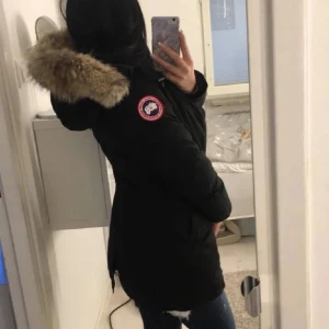 Svart canada goose med pälsdetalj - Säljer en svart parkajacka från Canada Goose med en mysig pälsdetalj på huvan. Jackan har en klassisk design med lång ärm och är perfekt för kalla dagar. Den har en snygg passform och är både stilren och funktionell.