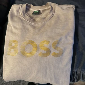 Beige och svart tröja från Hugo Boss - Säljer två stilrena tröjor från Hugo Boss. Den ena är beige med guldtext och den andra är svart med subtil logga. Perfekta för en avslappnad men snygg look. Passar bra till både jeans och chinos.