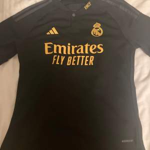 Säljer en svart Real Madrid fotbollströja från Adidas med guldfärgade detaljer och klubbmärke. Tröjan har korta ärmar och är i Aeroready-material för bra andningsförmåga. Perfekt för fans av laget! Sitter som xs-s. Köpt ny på unisport för 800