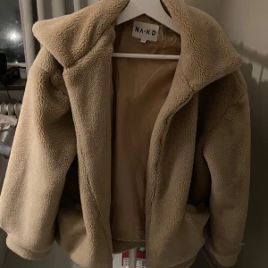 Beige teddyjacka från NA-KD - Mysig beige teddyjacka från NA-KD i storlek 38. Perfekt för kyliga höst- och vinterdagar. Super varm, haft på mig under snöstorm och överlevde😎🤘🏼Priset kan diskuteras.