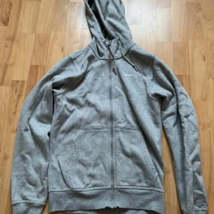 Grå hoodie från Nike - Säljer en grå hoodie från Nike i storlek M. Den är superbekväm och perfekt för både vardag och träning. Hoodien har en dragkedja framtill och en klassisk Nike-logga på bröstet. Den har långa ärmar och en praktisk huva med dragsko. Perfekt för höst och vår!