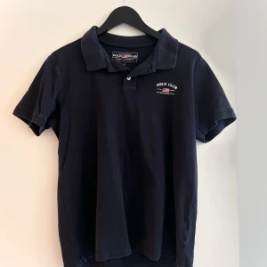 Polo Club marinblå piké t-shirt - En marinblå pikétröja med en liten amerikansk flagga på bröstet, klassisk polokrage och knappar i fram.   Storlek small. Mäter ca 47 cm över bröstet, se bild. Passar även medium och large.