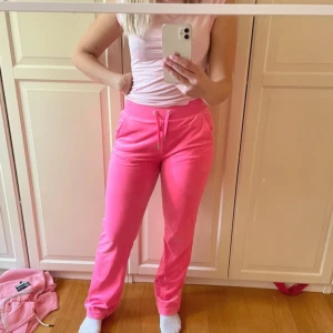 Rosa mjukisbyxor från Juicy Couture - Säljer ett par superbekväma rosa mjukisbyxor från Juicy Couture i storlek XS. Byxorna har en snygg snörning i midjan och är perfekta för en avslappnad dag hemma eller en chill dag ute. De är gjorda av mjukt material som känns skönt mot huden. Perfekt för dig som älskar en poppig färg i garderoben! 💖