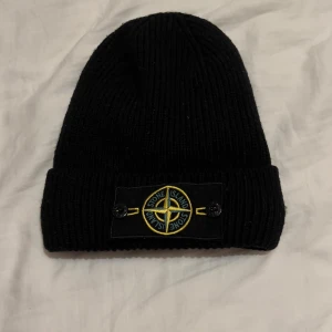 Svart ribbad mössa från Stone Island - Säljer en snygg svart ribbad mössa från Stone Island. Den har en broderad logga framtill i gult och grönt. Perfekt för att hålla sig varm och stilren under de kallare månaderna. 
