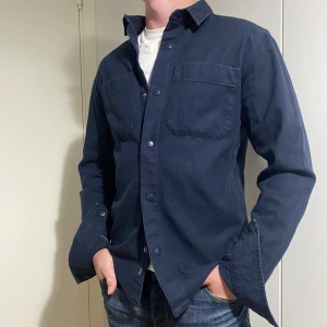 Mörkblå overshirt - Säljer en snygg mörkblå overshirt. Perfekt för höst och vår, med långa ärmar och knappar framtill. Den har två praktiska bröstfickor och en stilren design som funkar både till vardags och fest. Passar bra över en t-shirt för en avslappnad look.