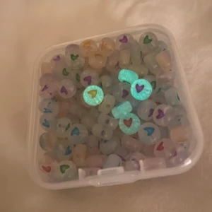 Lysande  pärlor med hjärtmönster - Säljer en ask med lysande pärlor som har små hjärtan i olika färger. Perfekta för att göra egna smycken eller pysselprojekt. Pärlorna lyser i mörkret och har en genomskinlig bas med hjärtan i färger som röd, blå, grön, lila och gul. Superkul för kreativa projekt eller som en unik detalj i smycken! De aktiveras genom att lysa med lampa på dem, 