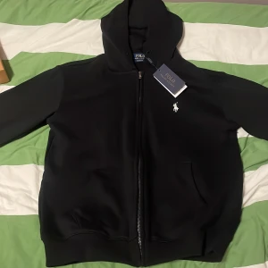 Svart hoodie från Polo Ralph Lauren  - OBS: snörena finns kvar jag har bara tagit ut dom för gillar ej snören på mina hoodies. Säljer en svart hoodie från Polo Ralph Lauren i nytt skick. Den har en dragkedja framtill och en liten vit logga på bröstet. Perfekt för höst och vår, med en skön passform och huva för extra mysfaktor. Passar både till vardags och lite mer avslappnade tillfällen. priset kan diskuteras och är ej hugget i sten 