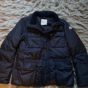 Moncler Field Jacket - Säljer en snygg marinblå Moncler jacka i mycket bra skick. Den har en quiltad design med dragkedja och knappar, samt flera praktiska fickor. Perfekt för höst och vinter! Säljer den för lite billigare pris för att på dragkedjan har en sak lossnat. Storlek 3 som motsvarar storlek M✅.Köpt på NK i Göteborg, NK påsen följer med och digitalt kvitto kan hittas vid förfrågan✅Priser är inte hugget i sten 🪨 Bara att skriva vid minsta fundering!