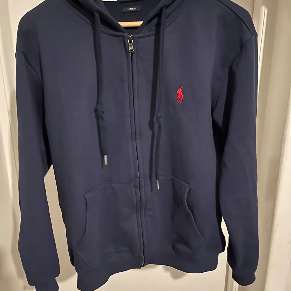 Säljer en snygg mörkblå zipup hoodie från Polo Ralph Lauren i storlek M. Den har en dragkedja framtill och en röd broderad logga på bröstet. Perfekt för både höst och vår, med en skön passform och fickor . Passar till alla tillfällen! Är helt ny o ren då den bara användes en enda gång. . Tröjor & Koftor.