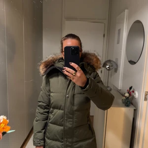 Olivgrön dunjacka från Hollies - Säljer en olivgrön dunjacka från Hollies i storlek 34. Jackan har en mysig pälskantad huva och stängs med dragkedja och knappar. Pälsdelen går att ta bort! Perfekt för kalla vinterdagar! 🧥❄️ Nypris ca 6000:-