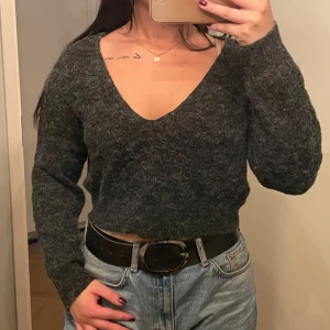 Mörkgrå stickad croppad tröja - Säljer en supermysig mörkgrå stickad tröja med v-ringning. Den är croppad och har långa ärmar, perfekt för höst och vinter. Tröjan är i bra skick och passar perfekt till jeans eller kjol. Perfekt för en avslappnad men stilren look!