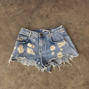 Blå jeansshorts - Säljer ett par coola blå jeansshorts med slitningar och fransiga kanter. Perfekta för en avslappnad sommarlook. De har en knapp och dragkedja framtill samt fem fickor. Passar perfekt för festivaler eller en dag på stranden!