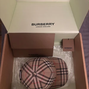 keps från Burberry - Säljer en snygg keps från Burberry i klassiskt beige, svart och rött mönster. Kepsen är i bra skick och har en justerbar rem baktill för perfekt passform. Perfekt accessoar för att lyfta vilken outfit som helst!!!  Tar endas mot byten med kvitto och box!