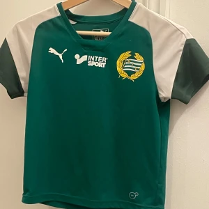 Grön fotbollströja och shorts från Puma och Craft - Säljer en grön fotbollströja från Puma med korta ärmar och vit logga, samt matchande gröna shorts från Craft med VW-logga. Tröjan har en klubbemblem på bröstet och är i mycket bra skick. Perfekt för unga fotbollsfans! Storlek 140, passar 9-10 år.