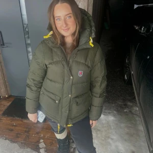  Fjällräven jacka expedition lite - Snygg och varm grön dunjacka från Fjällräven med huva och gula detaljer. Jackan har en dragkedja och knappar framtill samt praktiska fickor med lock. Perfekt för kyliga dagar. En pytte defekt som ej syns men är så populär och slutsåld överallt. Dam S