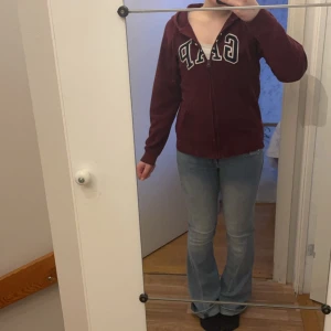Vinröd hoodie från GAP - Säljer en vinröd hoodie från GAP med dragkedja och stor logga på framsidan. Den har långa ärmar och en bekväm passform. Säljer då den har blivit lite kort i ärmarna❣️