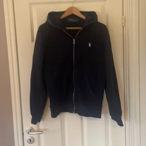 Mörkblå hoodie från Polo Ralph Lauren - Säljer en stilren mörkblå hoodie från Polo Ralph Lauren med dragkedja och huva. Tröjan har ett litet broderat märke på bröstet och är perfekt för en avslappnad look. Passar bra till både jeans och joggers.