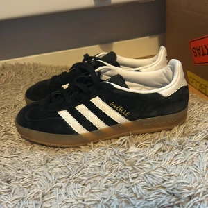 Svarta Adidas Gazelle sneakers - Snygga svarta Adidas Gazelle sneakers med vita detaljer och klassiska tre ränder på sidan. Skorna har en stilren design med snörning och en bekväm sula. Perfekta för en avslappnad stil.