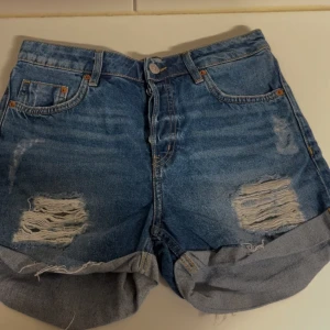 Blå jeansshorts med slitningar - Snygga blå jeansshorts med slitningar och upprullade kanter. De har en knappgylf och klassiska femficksdesign. Perfekta för en avslappnad stil.