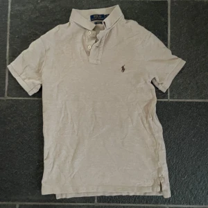 Beige kortärmad pikétröja från Ralph Lauren - Säljer en stilren beige pikétröja från Ralph Lauren med korta ärmar och klassisk krage. Tröjan har knappar framtill och den ikoniska broderade loggan på bröstet. Perfekt för en avslappnad men snygg look.