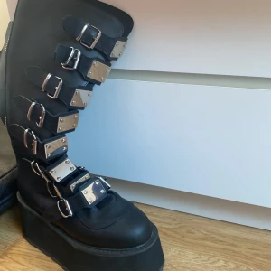 Damned-318 Boots - Snygga svarta boots från Damned med flera spännen och en dragkedja på bamsidan. De har en hög platåsula och är tillverkade i syntetmaterial. Perfekta för en edgy look. 