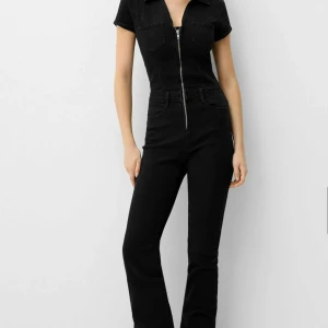 Svart jumpsuit med dragkedja - Snygg svart jumpsuit med korta ärmar och dragkedja framtill. Den har en krage och två bröstfickor😇Säljer pga förliten storlek,aldrig använd med prislapp kvar