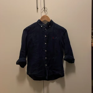 Mörkblå skjorta med knappdetaljer - Snygg mörkblå skjorta från Ralph Lauren med klassisk krage och knappar framtill. Den har långa ärmar och en liten broderad logga på bröstet. Perfekt för en stilren look.