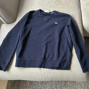 Mörkblå sweatshirt från Lacoste - Säljer en stilren mörkblå sweatshirt från Lacoste med det klassiska krokodilmärket på bröstet. Tröjan har långa ärmar och en rund halsringning. Perfekt för en avslappnad look. Hör av dig vid intresse!
