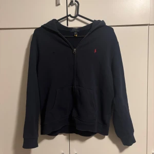 Mörkblå hoodie från Ralph Lauren - Säljer en snygg mörkblå hoodie från Ralph Lauren med dragkedja och det klassiska röda logotypen på bröstet. Den är i använt skick och har ett litet litet hål på vänstra bröst men inget som syns vid användning. Skriv för fler bilder elr funderingar 👍