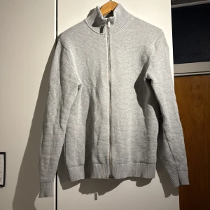 Grå stickad kofta från Selected Homme - Säljer en stilren grå stickad kofta från Selected Homme. Koftan har en dragkedja framtill och en hög krage som ger extra värme. Perfekt för kyligare dagar. Passar bra till både jeans och chinos. Perfekt nu till våren. Skick 7/10, tröjan är sparsamt använd och inga större tecken på användning 